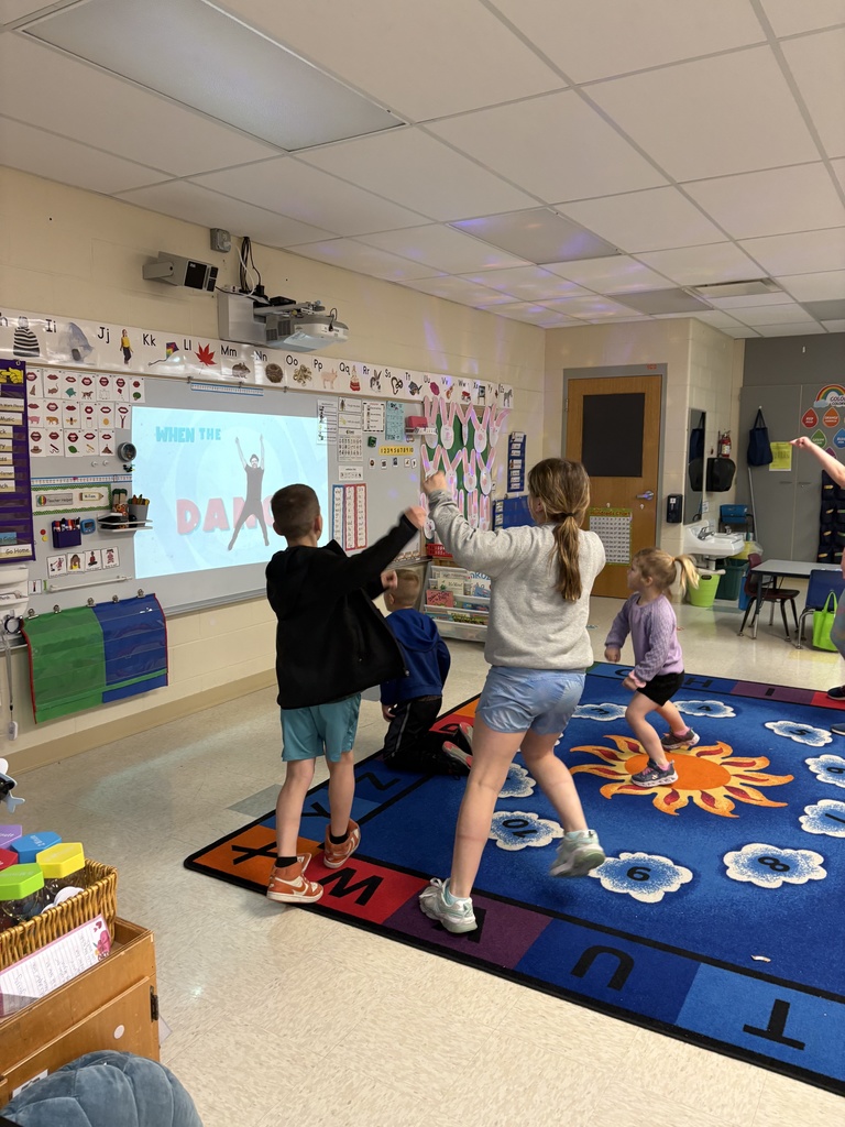 Children dance and move on a classroom rug while following a projected activity on the wall.