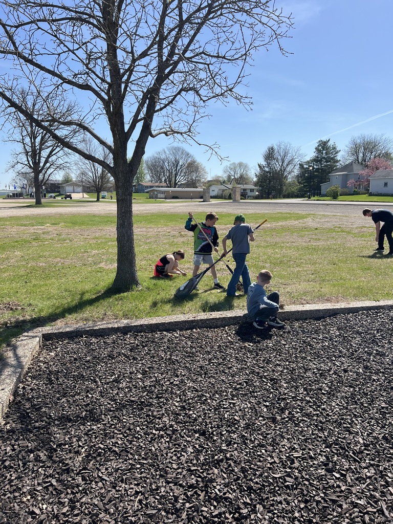 Children play and dig in a grassy park near a tree on a sunny day, with one adult supervising in the background.