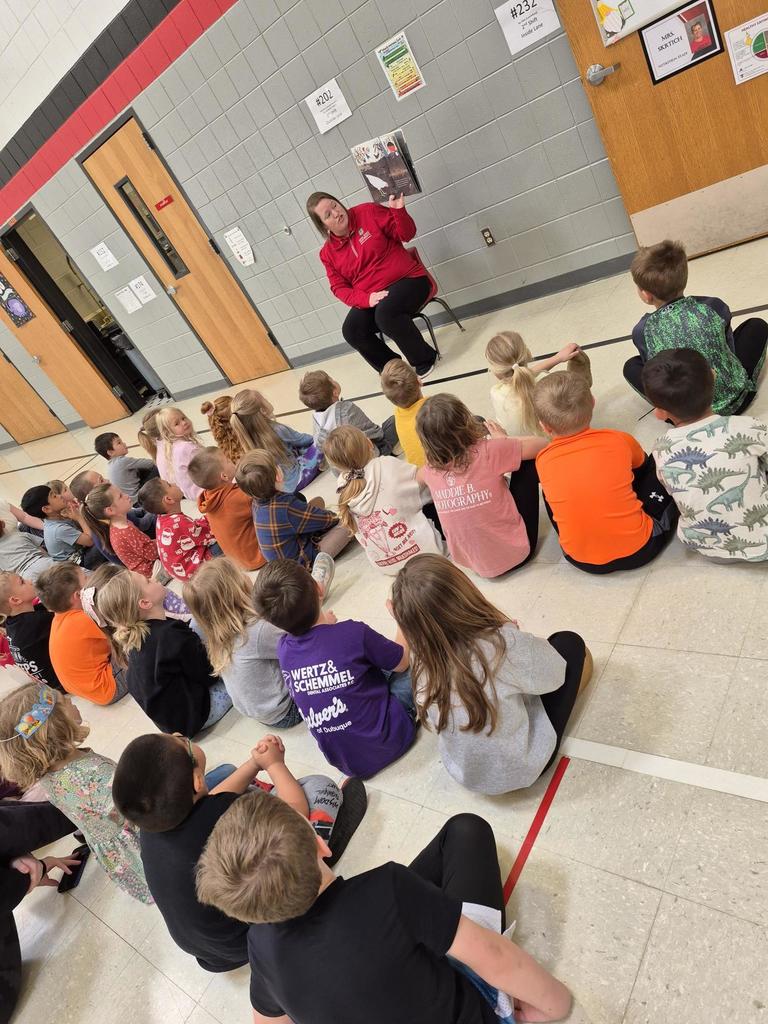 Children sit on a classroom floor facing a teacher in a red shirt, who reads a book aloud during group story time.