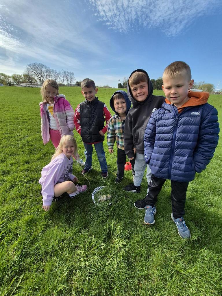 Six children crouch close together outdoors, smiling at the camera beside a small pile of grass and flowers on a plate.