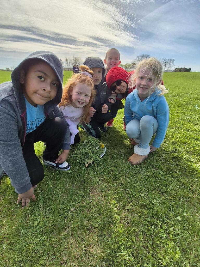 Six children crouch close together outdoors, smiling at the camera beside a small pile of grass and flowers on a plate.