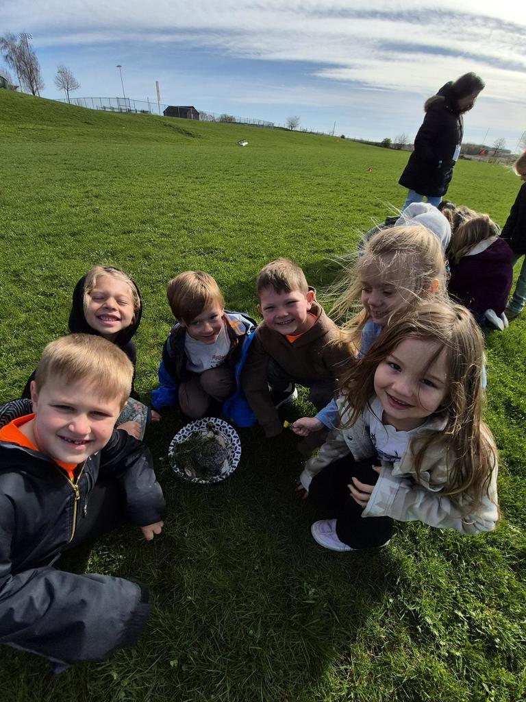 Six children stand and kneel in a grassy field, gathered around a small plate of natural materials, smiling in bright sunlight