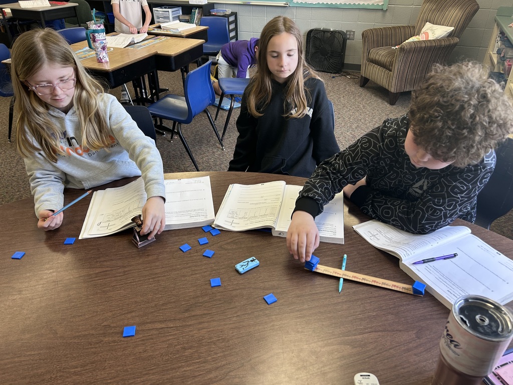 Three students sit at a classroom table using rulers and blue tiles to solve math problems, with open workbooks and pencils nearby.
