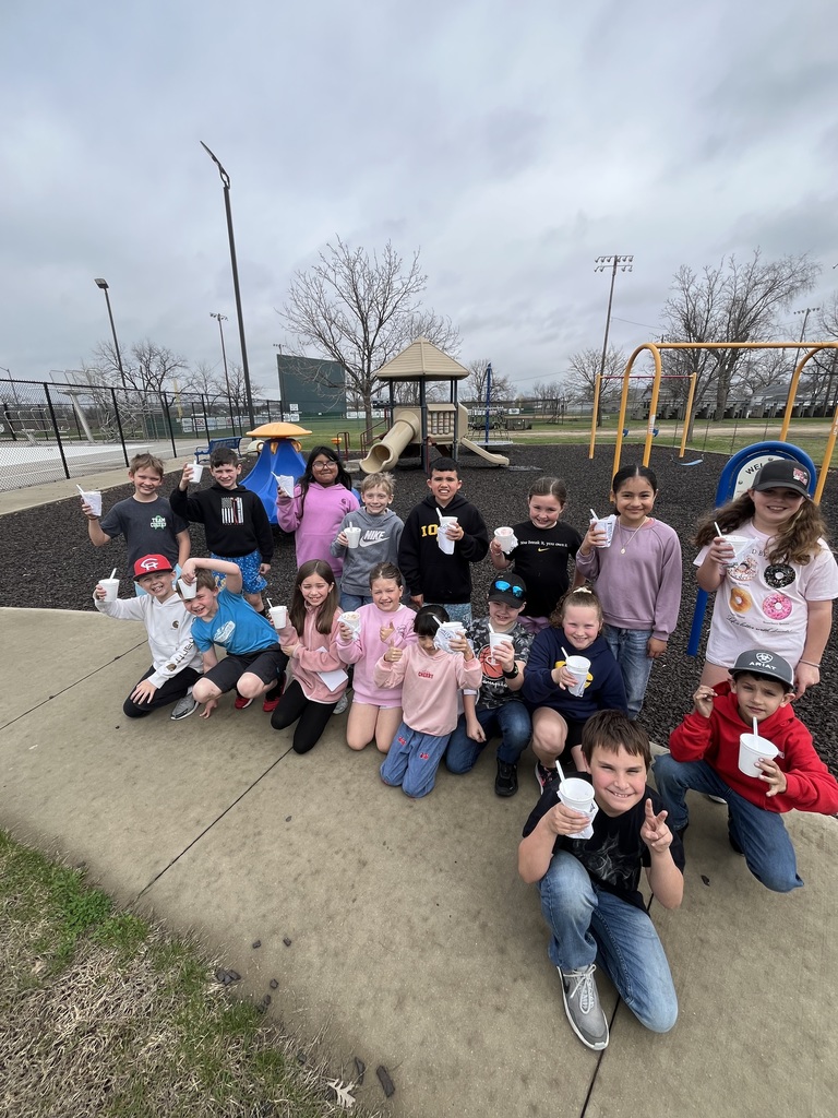 Group of elementary students pose on a playground, smiling and holding cups, with slides and swings visible behind them on a cloudy day.