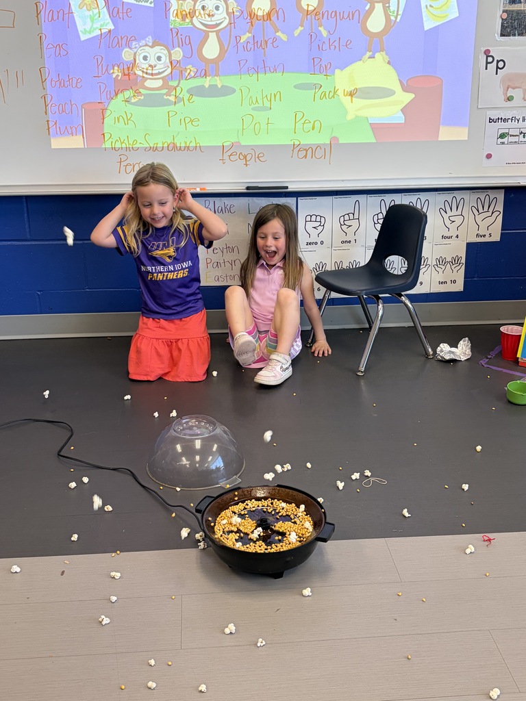 Two young girls sit on a classroom floor, smiling as popcorn pops from a small machine, scattering kernels around them.