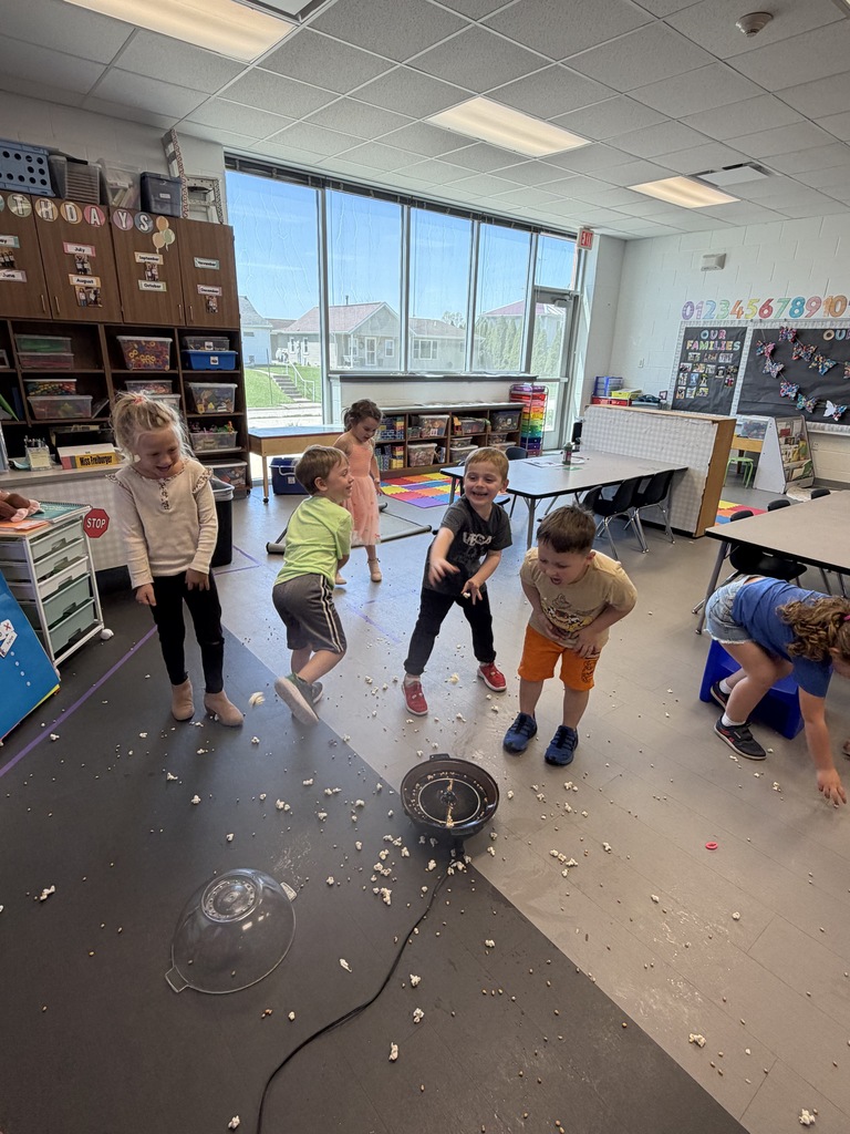 Kids in a classroom laugh and jump around a popcorn popper as kernels scatter across the floor during a playful experiment.