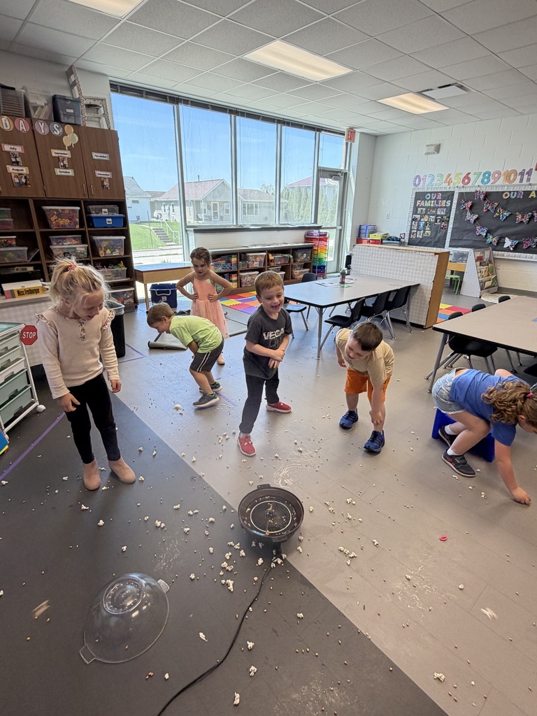Children in a classroom laugh and move around as popcorn pops onto the floor from a small machine during a hands-on activity.