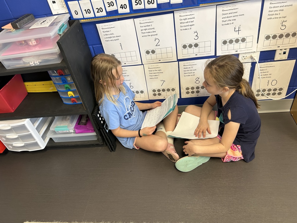 Two girls sit on the floor by a classroom wall, taking turns reading their writing to each other during partner work.