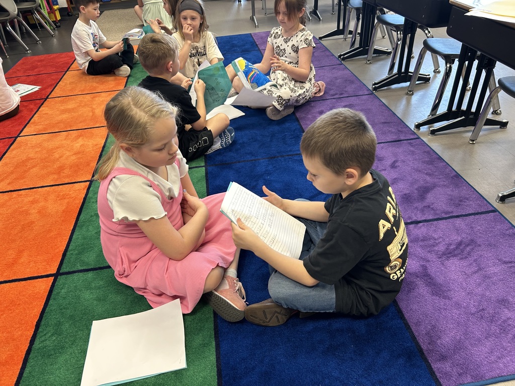 Students sit in pairs on a colorful rug, reading and discussing their writing with partners in a classroom.