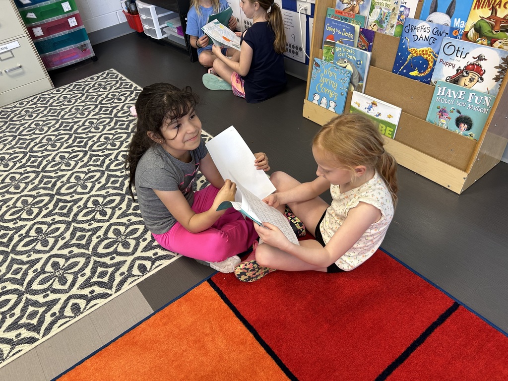 Two young students sit on a classroom rug, smiling and sharing their writing while other pairs work nearby.