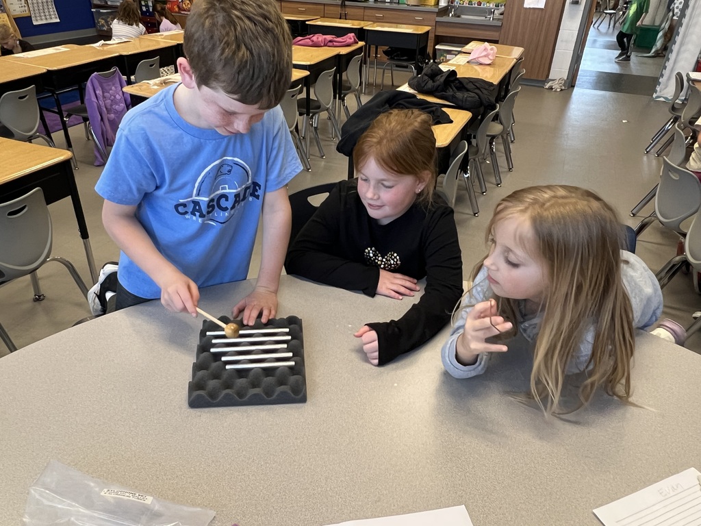 Three students gather around a table, watching as one taps metal bars with a mallet during a classroom experiment.