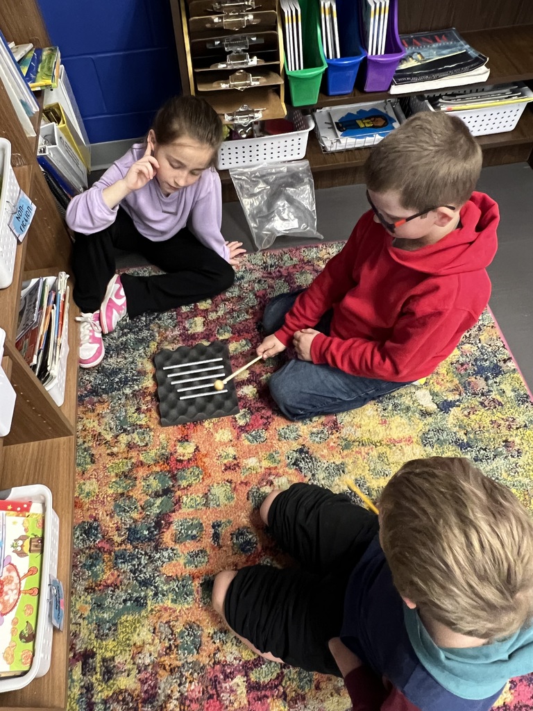 Two students sit on a rug, one tapping metal bars with a mallet while the other listens and observes closely.