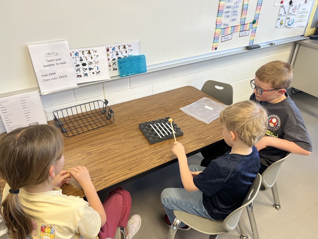 Three students sit at a table, taking turns tapping metal bars with a mallet during a hands-on sound activity.