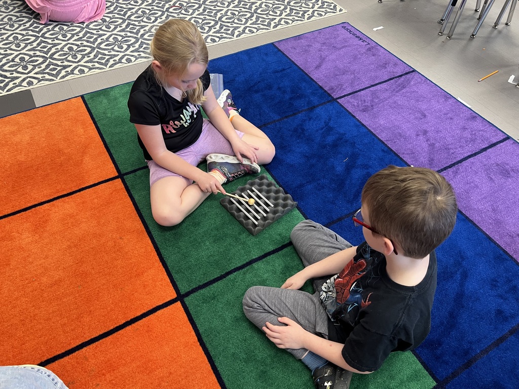 Two students sit on a rug, one tapping metal bars with a mallet while the other watches during a sound activity.