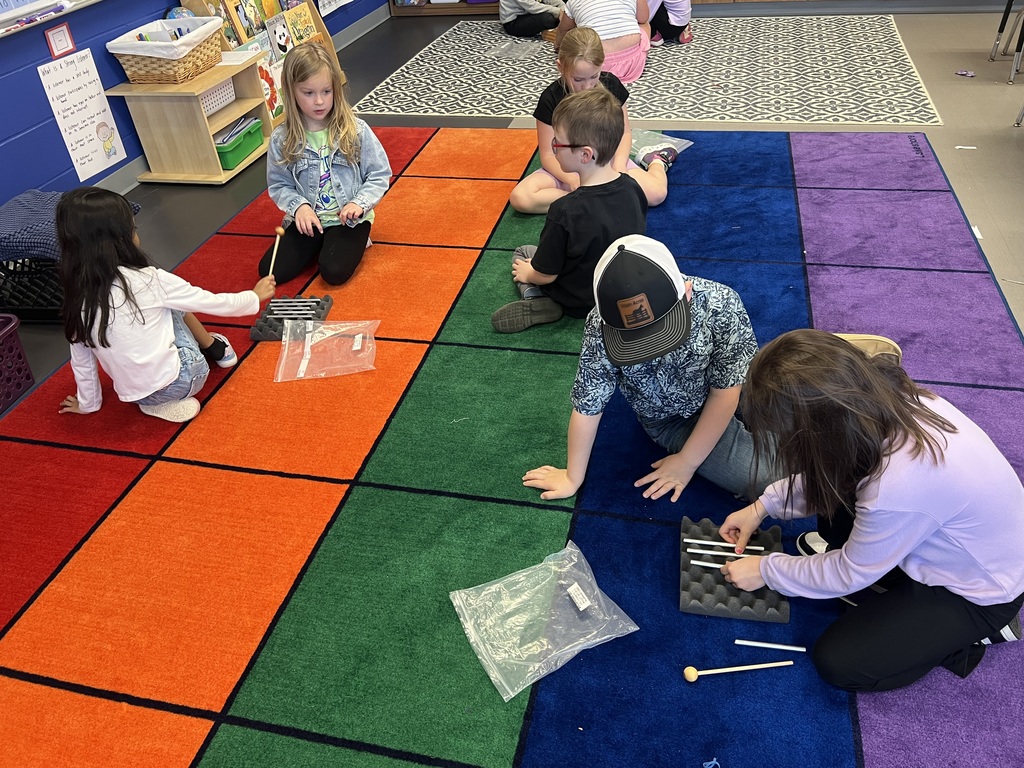 Small groups of students sit on a colorful rug, taking turns tapping metal bars and exploring sound together.