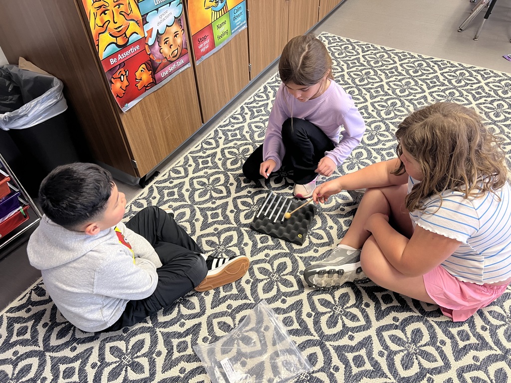 Three students sit on a patterned rug, taking turns tapping metal bars with a mallet in a small group activity.