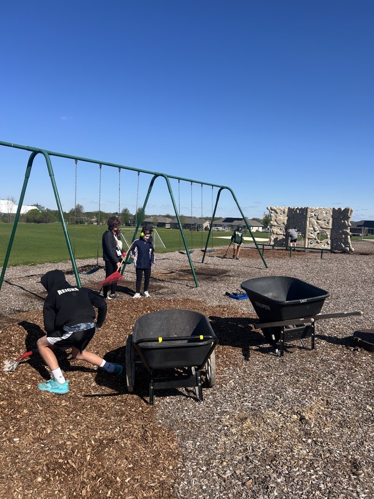 Students rake and spread wood chips around a playground with swings and climbing equipment in the background.