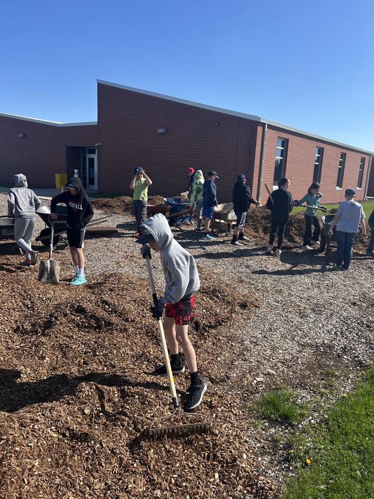 Students spread wood chips with rakes and shovels outside a brick school building on a sunny day.