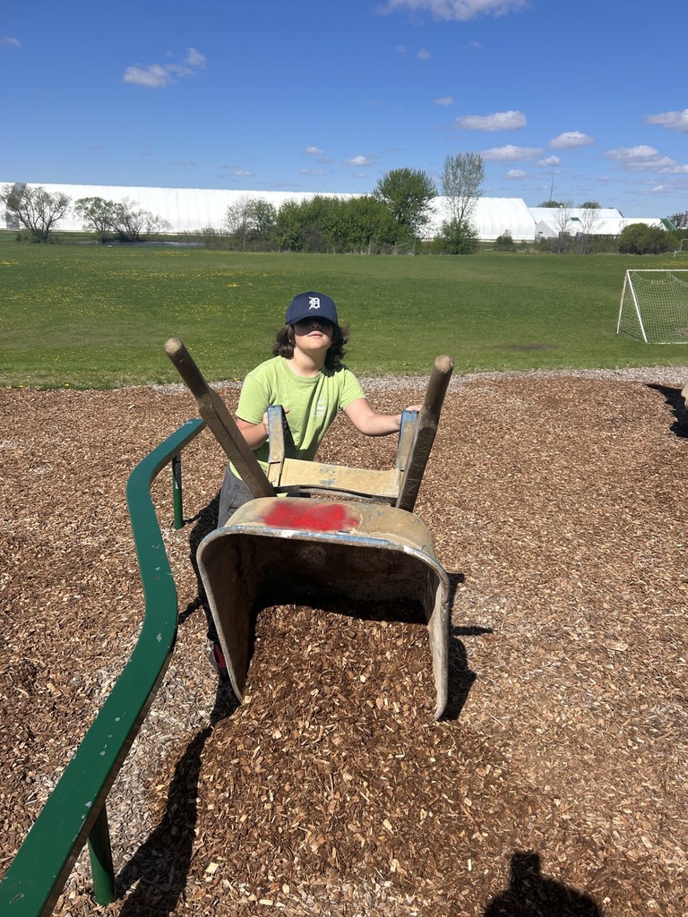 Student tips a wheelbarrow to dump wood chips onto the ground in a grassy playground area.
