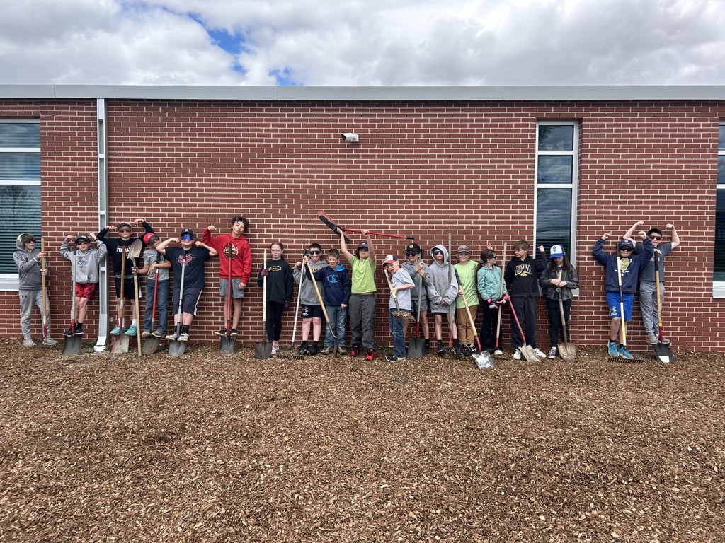 Group of students lined up against a brick wall holding shovels and rakes after outdoor cleanup work.