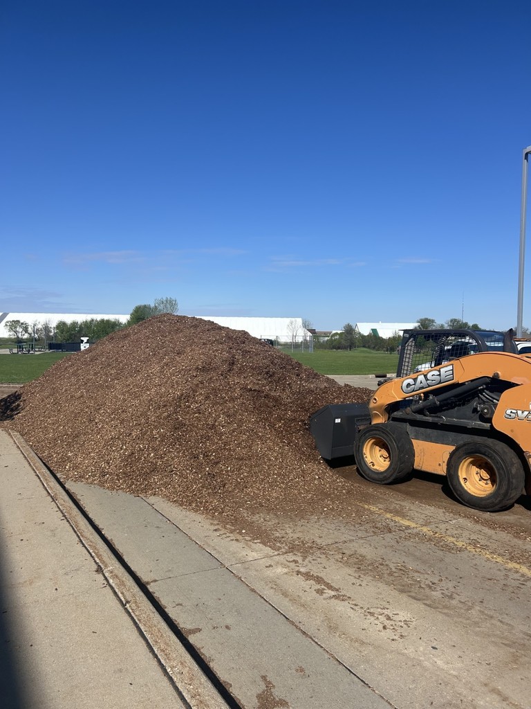 Large pile of wood chips sits near a sidewalk beside a skid steer loader on a sunny day.