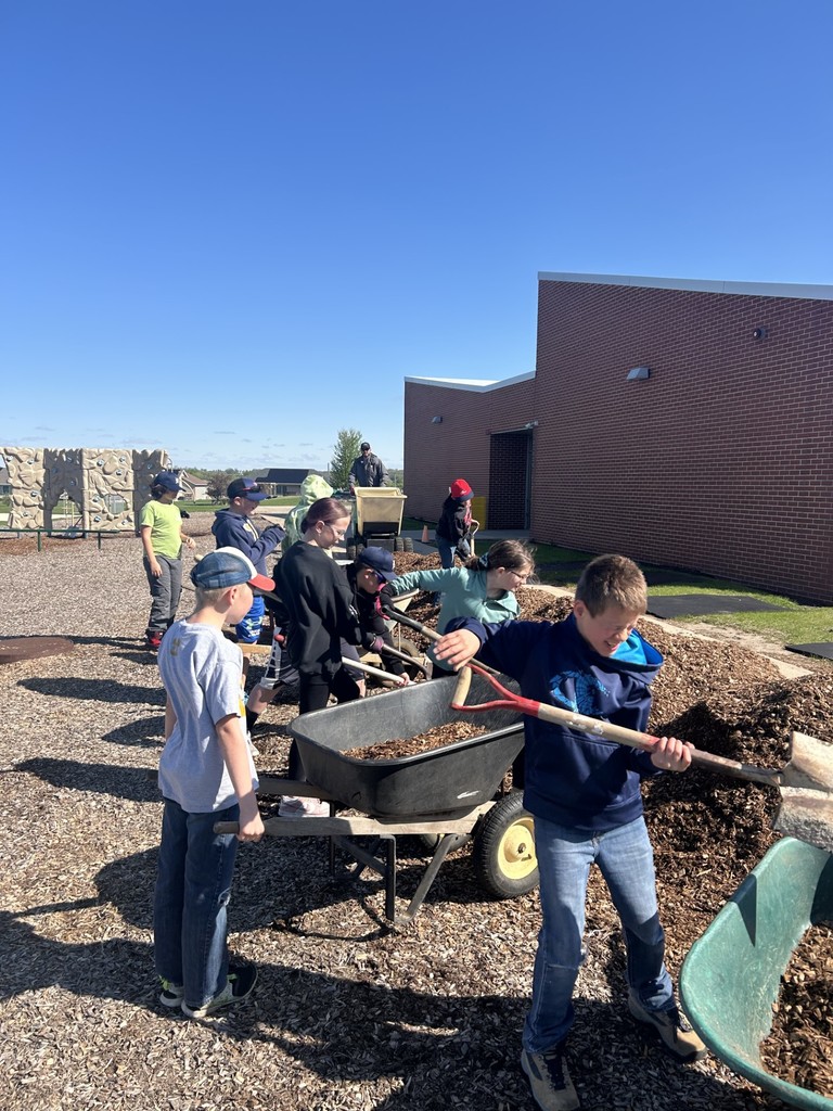 Students shovel and move wood chips into a wheelbarrow near a school building under a clear blue sky.