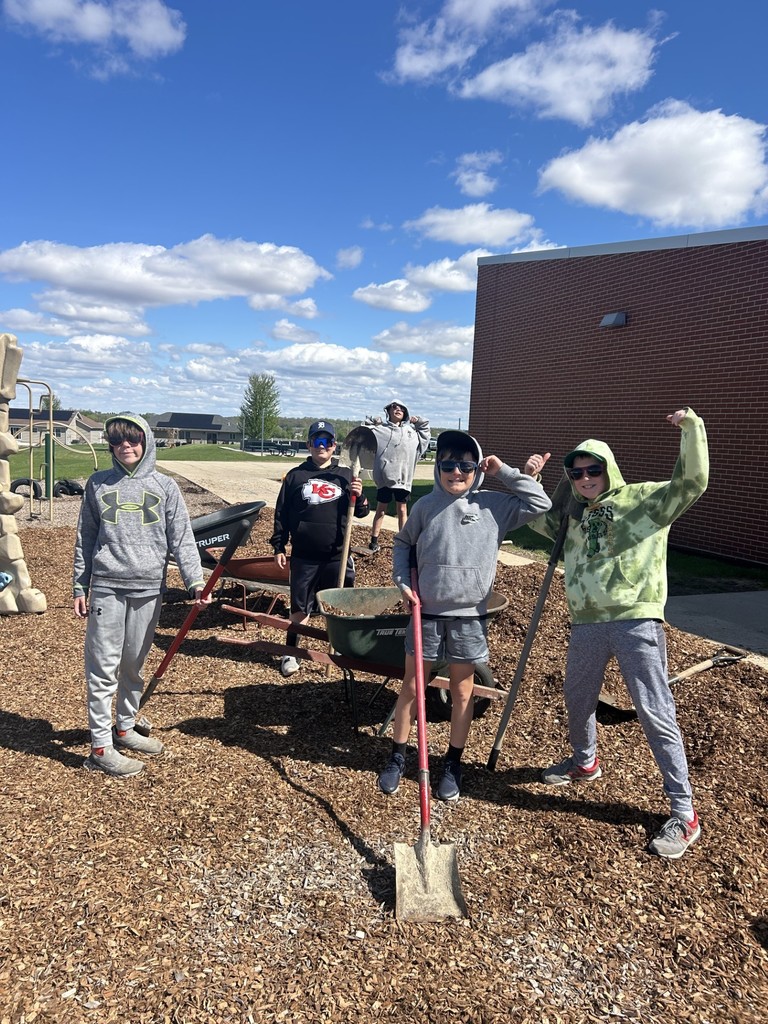 Students pose with shovels and a wheelbarrow, smiling and flexing after working with wood chips outdoors.