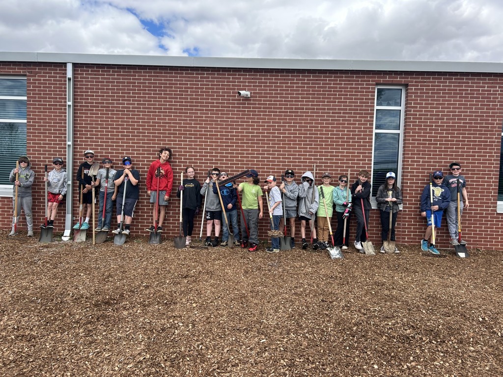 Group of students lined up against a brick wall holding shovels and rakes after outdoor cleanup work.