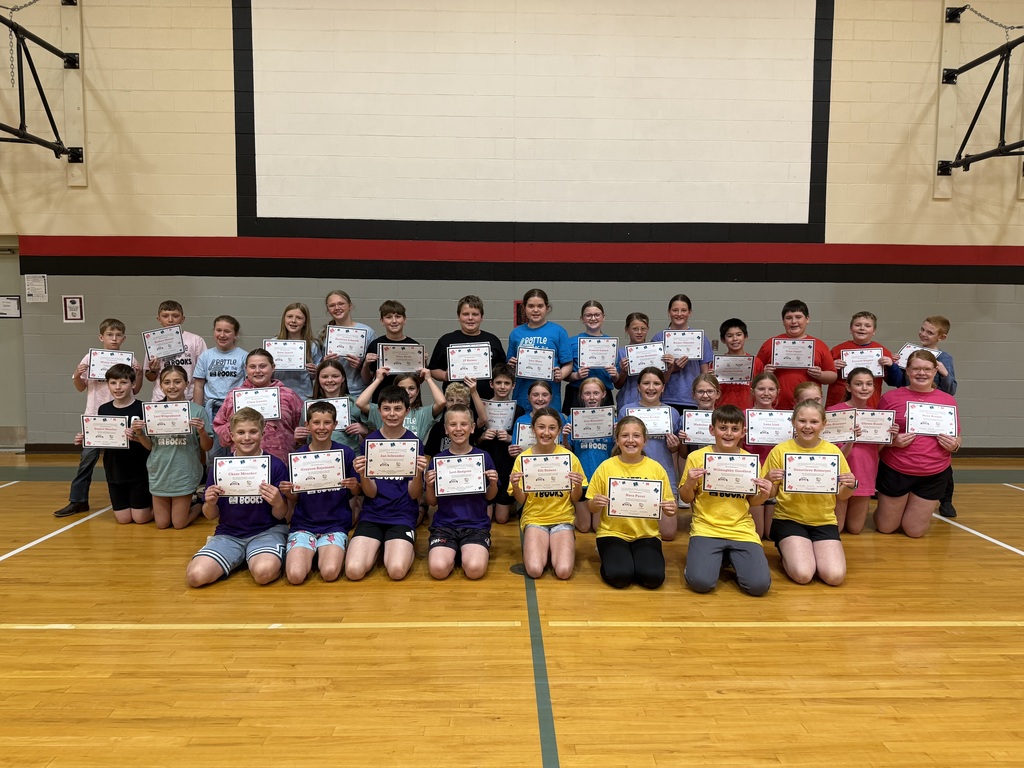 Group of students in a gym kneel and stand in rows, smiling and holding certificates against a wall with red and black stripes.