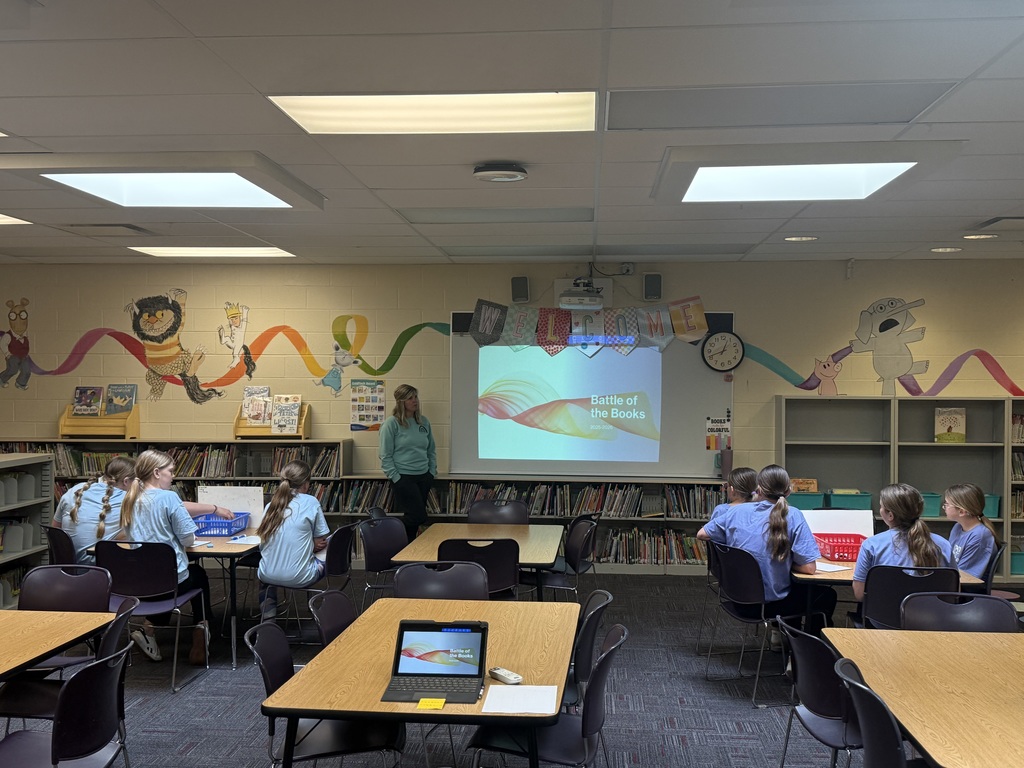 Students sit at tables in a library while a teacher stands near a screen displaying “Battle of the Books” under a welcome banner.