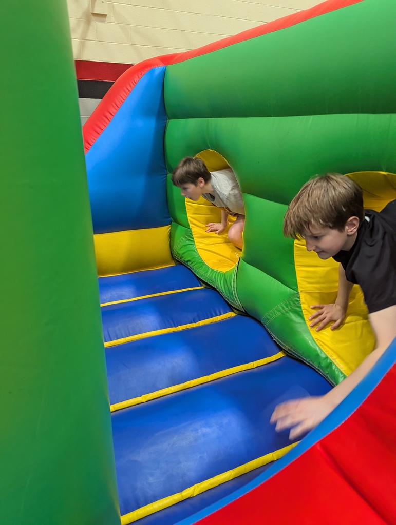 Two children crawl through round openings inside a colorful inflatable obstacle course with green walls and a blue padded floor.