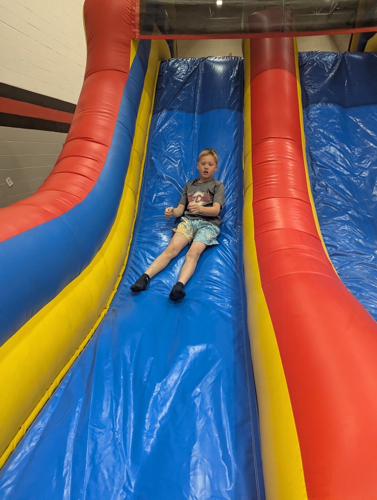 Child slides down a large inflatable slide in a gym, seated on a bright blue lane with red and yellow sides.
