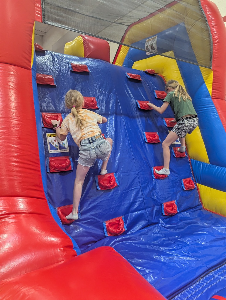 Two children climb an inflatable wall with red grips, surrounded by bright red, yellow, and blue padded sides in a gym.