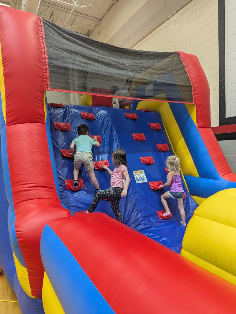 Three young children climb a steep inflatable wall using red footholds on a blue surface inside a gym play structure.