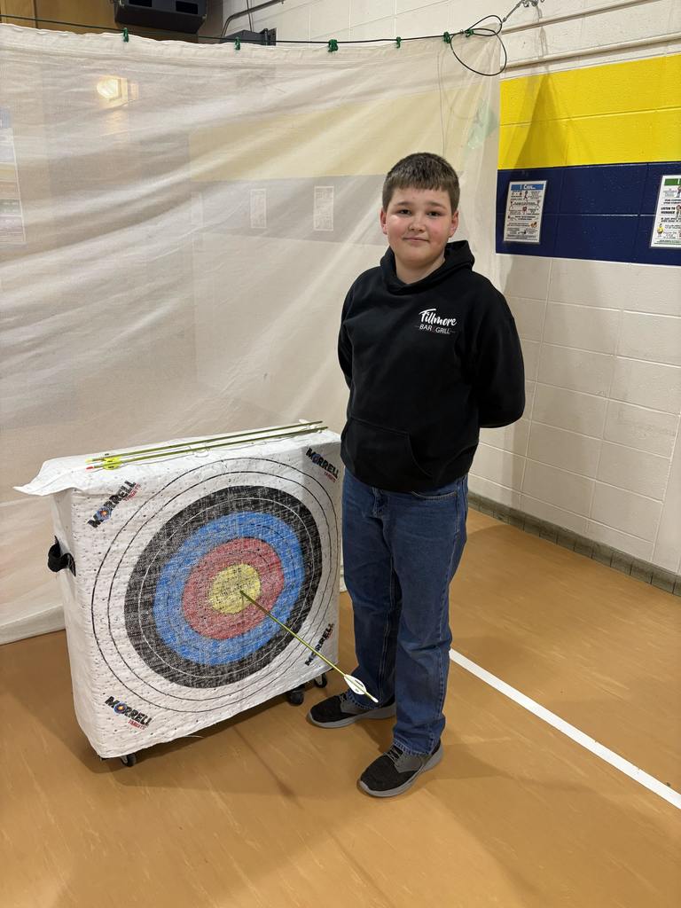 Boy in black hoodie stands next to archery target with an arrow in the bullseye, hands behind his back.