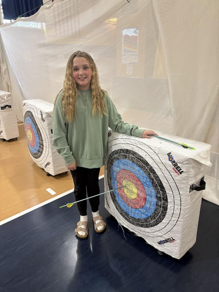 Girl in green sweatshirt stands beside target, smiling and pointing to an arrow in the center bullseye.