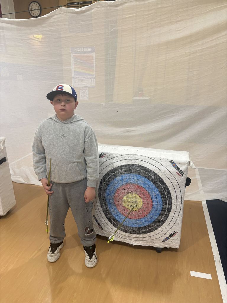 Boy in gray hoodie and cap stands next to target with an arrow in the bullseye, holding additional arrows.