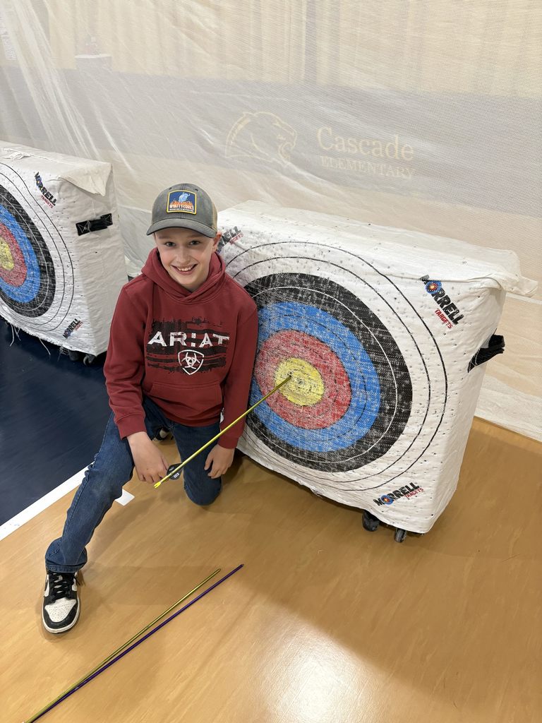 Boy in red hoodie kneels by archery target, grinning and pointing to a bullseye shot with arrows on the floor nearby.