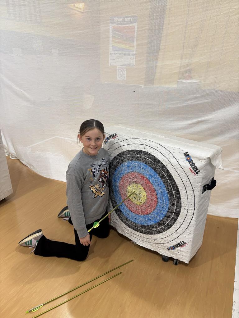 Girl kneels beside archery target, smiling and pointing to an arrow lodged in the yellow center.