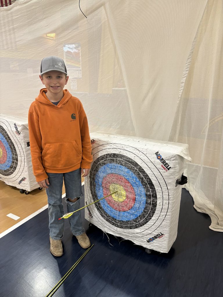 Boy in orange hoodie and cap stands by target, pointing to a bullseye hit; extra arrows lie on the gym floor.