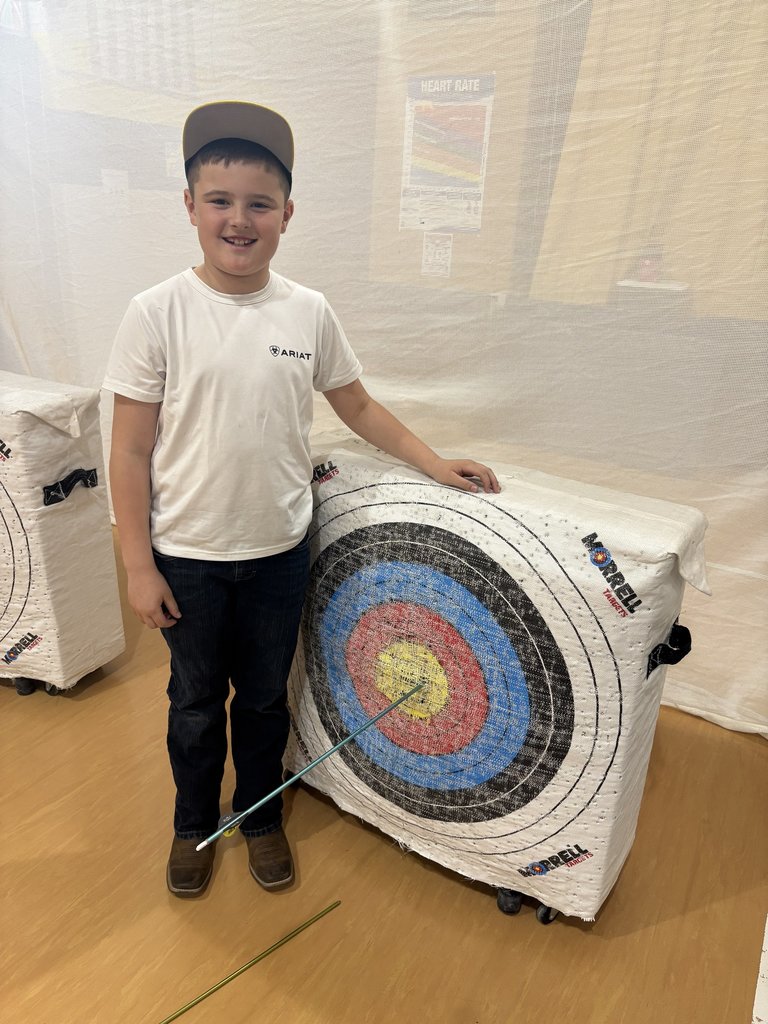 Boy in white shirt and cap smiles beside archery target, pointing to an arrow hitting the yellow bullseye in a gym.