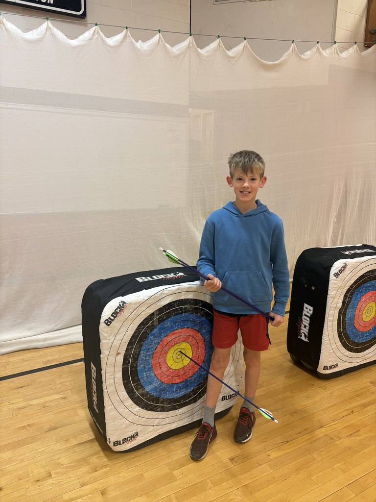 Boy in blue hoodie and red shorts stands by archery target, smiling and holding arrows, with one hitting the bullseye.