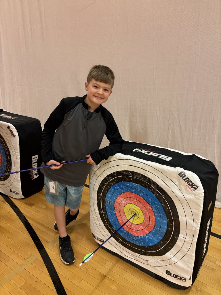 Boy leans on archery target, smiling and pointing to an arrow hitting the yellow bullseye in the gym.