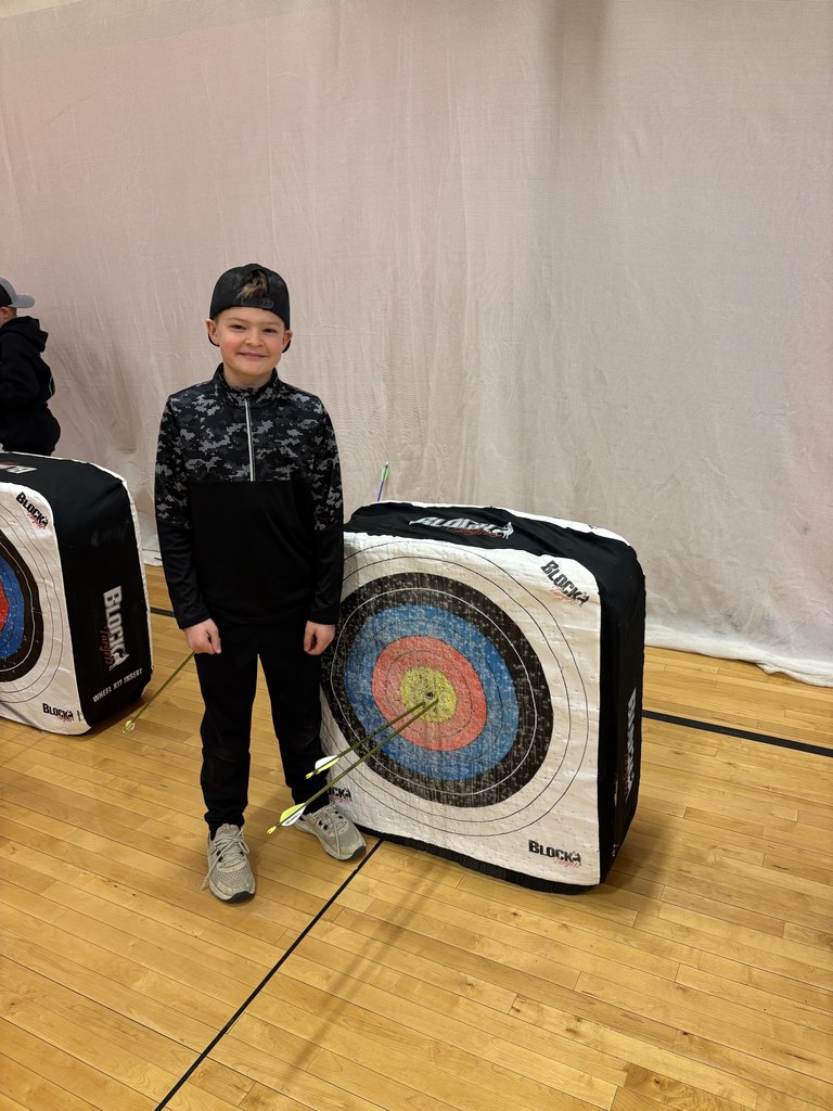 Boy in black camo top and cap stands by target with multiple arrows clustered near the center rings.