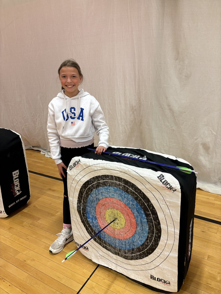 Girl in white “USA” hoodie smiles next to target, pointing to an arrow in the bullseye on a gym floor.