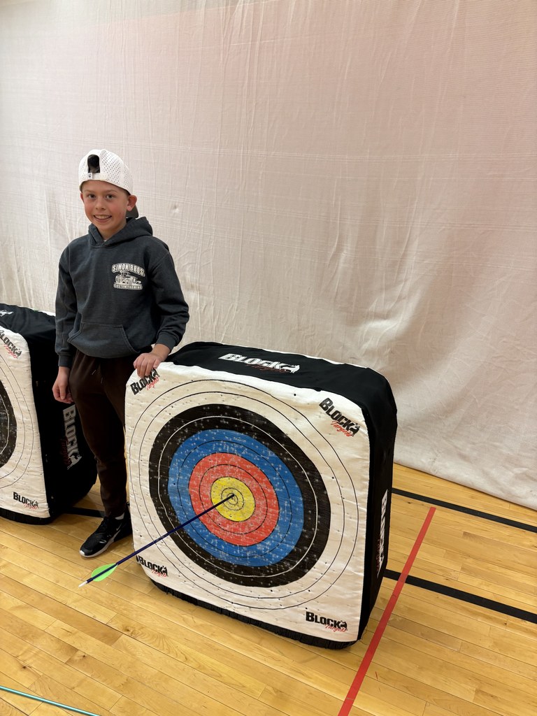 Boy in gray hoodie and white cap stands beside target, smiling as an arrow lands in the yellow center.