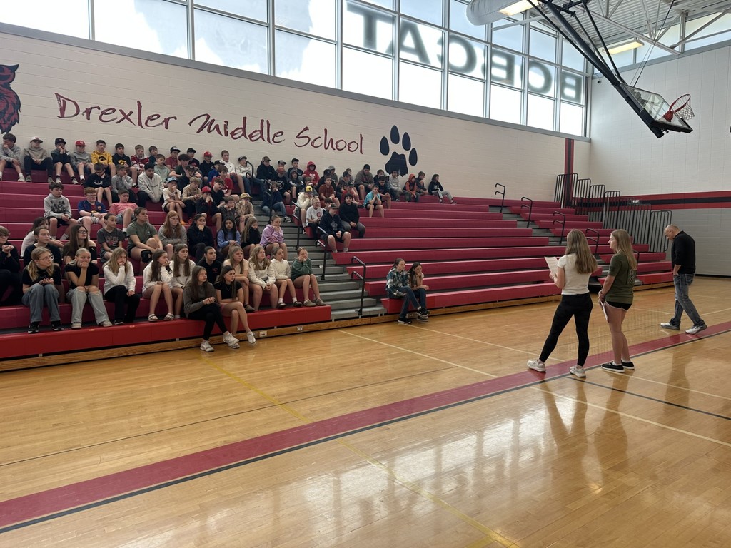 Students seated in Drexler Middle School gym bleachers while two students speak at the front of the assembly.