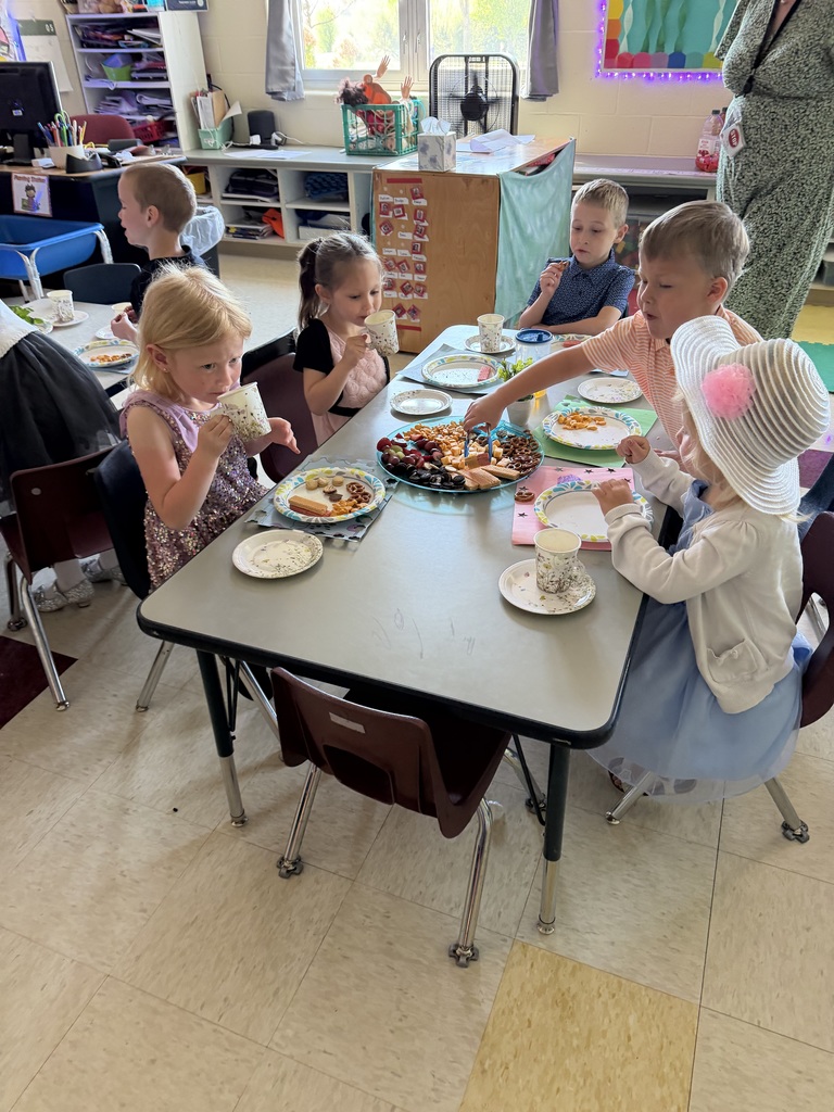 Kindergarten students and teachers dressed in spring hats enjoy a classroom tea party with snacks, tea cups, and colorful decorations.