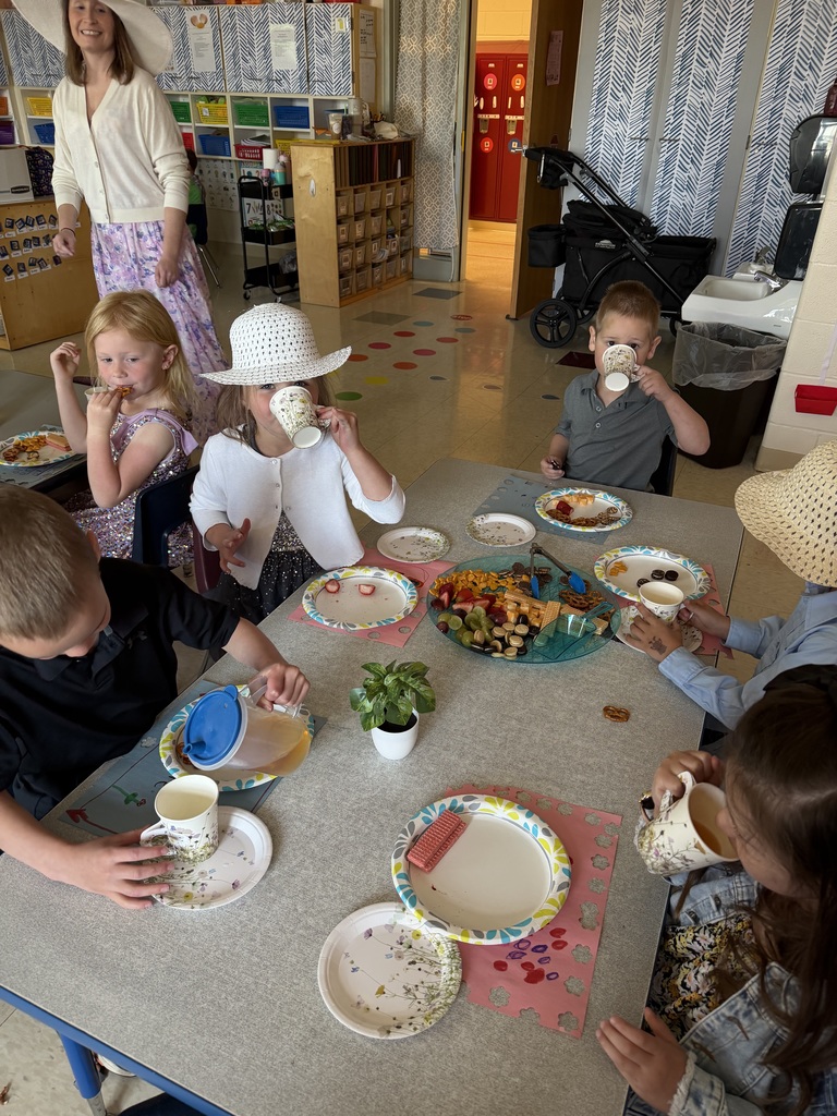 Kindergarten students and teachers dressed in spring hats enjoy a classroom tea party with snacks, tea cups, and colorful decorations.