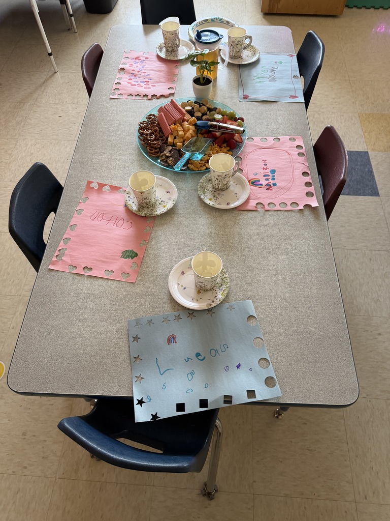 Kindergarten students and teachers dressed in spring hats enjoy a classroom tea party with snacks, tea cups, and colorful decorations.